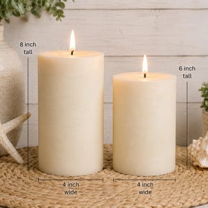 May include: Two cylindrical off-white pillar candles with lit flames. The left candle is 8 inches tall and 4 inches wide. The right candle is 6 inches tall and 4 inches wide. Both candles have a smooth wax surface and are positioned upright.