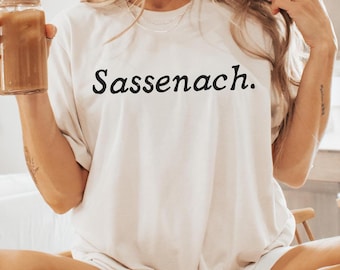 May include: A white t-shirt with the word "Sassenach." printed in black on the front. The person wearing the shirt is sitting on a counter with their legs crossed. They are wearing white socks with black stripes.