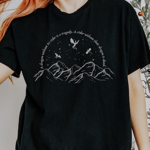 Op de afbeelding: Zwart T-shirt met een witte afbeelding van bergen, sterren en draken. De tekst op het T-shirt luidt "A dragon without its rider is a tragedy. A rider without their dragon is dead."