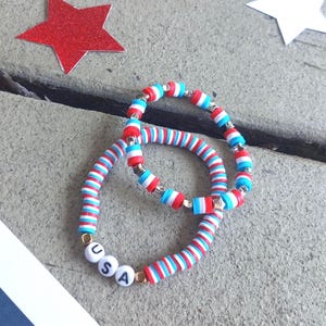 May include: Two stretch bracelets made of small, flat, circular clay beads in alternating red, white, and blue colors. One bracelet features white spherical beads with the black letters U, S, and A printed on them, accented by small gold-toned spacers.