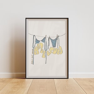 May include: A framed print with a hand-drawn illustration of the word "Happy" written in yellow letters with a blue bikini top hanging from each letter. The word "Sun" is written in yellow letters with a blue bikini bottom hanging from each letter. The text "Girls just wanna have fun" is written in small, yellow letters at the bottom left corner of the print.