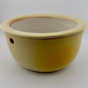 Op de afbeelding: Een gele keramische kom met een afgeronde vorm en een brede, lichtgele rand. De kom heeft een klein afvoergat aan de zijkant. De binnenkant is lichtbeige van kleur. Deze kom is geschikt voor planten of decoratieve doeleinden.