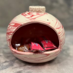 May include: A white ceramic candy dish shaped like a Christmas ornament with red marbling. The dish is filled with candy.