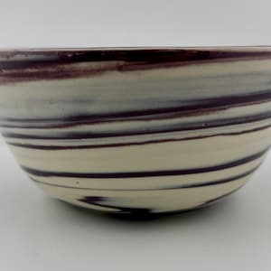 May include: A ceramic bowl with a cream-colored base and dark purple horizontal stripes. The rim of the bowl is a solid dark purple. The bowl has a wide, shallow shape, suitable for serving or display.