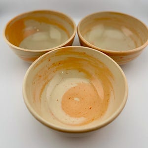 May include: Three ceramic bowls with a cream-colored interior and a swirl of orange and cream on the inside. The rims and outer surfaces of the bowls are orange. The bowls are arranged in a triangular pattern.