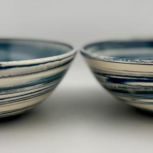 May include: Two ceramic bowls with a marbled design. The bowls have a white base with blue and gray swirls. The rims are a darker shade of blue. The bowls are round and appear to be handmade.