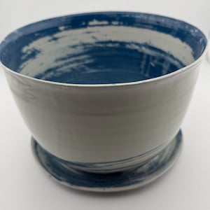 May include: A ceramic planter with a wide, open bowl shape. The interior and rim are painted with a deep blue color, contrasting with the off-white exterior. The planter sits on a matching saucer base, creating a cohesive design.