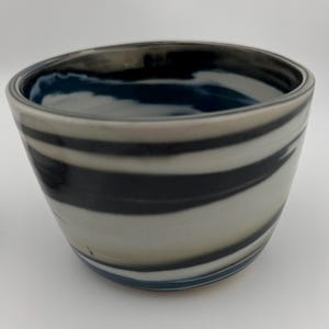 May include: A ceramic bowl with a unique design. The bowl features alternating bands of black, white, and gray, creating a striped pattern. The interior has a dark blue hue. The bowl's rim is slightly irregular, adding to its handmade aesthetic.