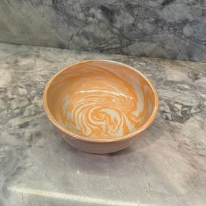 May include: A swirl-patterned ceramic bowl with a light orange and white glaze. The bowl is empty and sitting on a gray countertop.