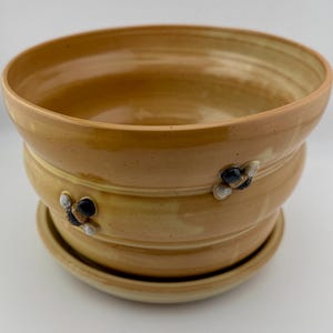 May include: A handcrafted, light brown ceramic planter with a matching saucer. The planter features a tiered design and is adorned with three-dimensional bee embellishments. The glazed finish gives it a glossy appearance, suitable for indoor or outdoor use.