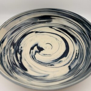 May include: A ceramic bowl with a cream-colored interior and a swirling navy blue pattern. The bowl's rim is a darker shade of blue, and the design creates a spiral effect. The bowl is round and appears to be handmade.