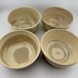 May include: Four beige ceramic bowls with a light brown swirl pattern inside. The bowls have a simple, rounded design and are arranged in a square formation. The bowls are likely for serving food.