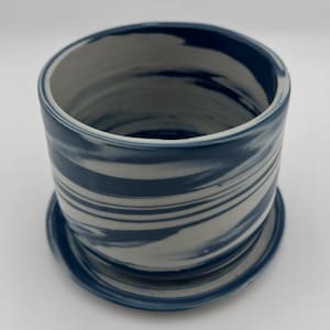 May include: A cylindrical ceramic planter with a matching saucer. The planter features a swirling pattern of blue and white glaze. The design is a marbled effect, with the colors blending together. The planter is suitable for small plants or succulents.