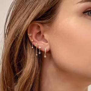 May include: Close-up of an ear displaying gold earrings with celestial motifs. The earrings incorporate crescent moons, stars, and dangling charms. The earrings are decorated with tiny, sparkling gemstones.