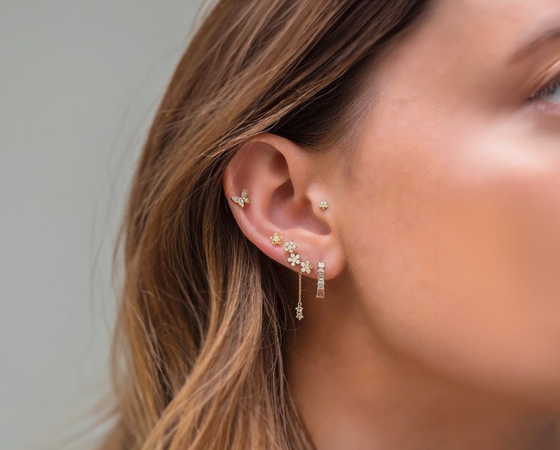 K&ouml;nnte beinhalten: Nahaufnahme eines Ohrs mit mehreren goldenen Ohrringen. Die Ohrringe zeigen einen Schmetterling, eine kleine Blume und eine gr&ouml;&szlig;ere Blume mit einer baumelnden Kette. Die Kette hat einen kleinen Sternanh&auml;nger am Ende.