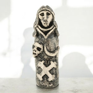 May include: A ceramic figurine of a woman with a skull and a cross on her chest. The figurine is painted in shades of grey and white.