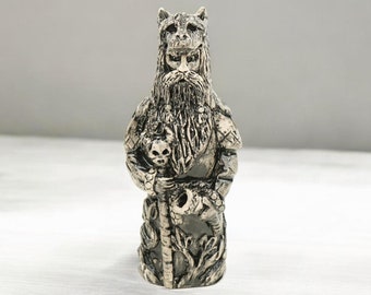 May include: A detailed silver figurine of a bearded man with a wolf-like head, holding a staff with a skull at the top. The figurine is standing on a white surface.