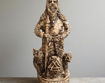 Puede incluir: Estatua beige de una figura barbuda con lanza y espada, flanqueada por dos lobos. La estatua tiene tallas detalladas y un aspecto desgastado. La base está inscrita con la palabra "Odin".