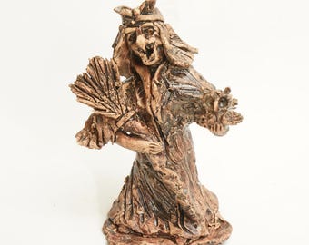 May include: A ceramic figurine of a witch with a broom and a flower in her hand. The figurine is painted in brown and black tones.