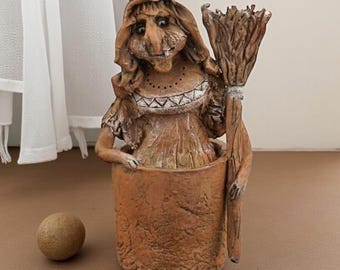 May include: A whimsical, brown ceramic witch figurine holding a broom, emerging from a cylindrical pot. The witch has a long nose, a headscarf, and a decorative collar. A small, round, brown ball sits beside the pot.