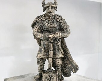 Puede incluir: Una estatua de cerámica de un guerrero vikingo sosteniendo un martillo. El guerrero lleva un casco con cuernos, una túnica de cota de malla y una capa. El martillo tiene un símbolo de runa en su cabeza.