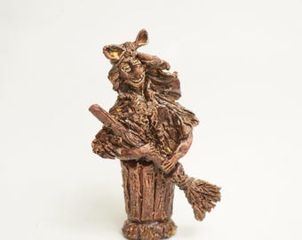 May include: A ceramic figurine of a witch with a brown and yellow painted finish. The witch is holding a broom and wearing a hat. The figurine is standing on a wooden base.