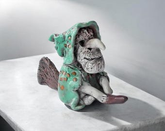 May include: A whimsical ceramic figurine of a seated creature with a long nose and a green hooded cloak adorned with orange spots. The figure has a textured, gray face and is perched on a brown, broom-like base.