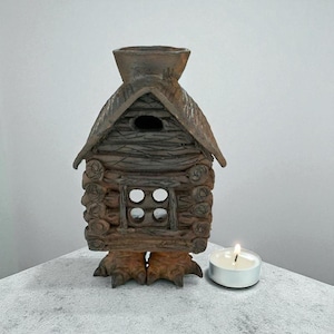 May include: A brown ceramic candle holder in the shape of a log cabin. The cabin features a small window with four panes and a chimney. A lit white tea light candle is placed next to the cabin, providing a warm light.