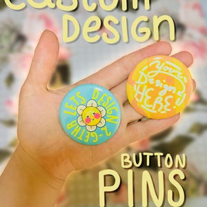 May include: Two custom design button pins are held in a hand. One is light blue with a smiling flower and the text "Lets Design 2-Gether". The other is yellow and orange with the text "Your Design HERE!!". The words "Custom Design" and "Button Pins" are also in the image.
