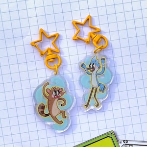 May include: Two yellow star-shaped keychains with cartoon characters. One keychain features a blue bird with a red beak and the other keychain features a brown raccoon.