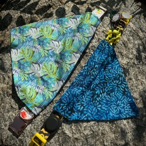 May include: Two dog bandanas with different patterns. One bandana is blue with a white floral pattern and the other is green and blue with a tropical leaf pattern. Both bandanas have yellow and black adjustable collars.