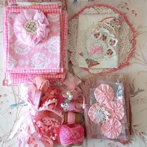 May include: A collection of craft supplies in shades of pink and white, including stacked fabric squares with a ruffled flower embellishment, lace doilies, pink ribbons, yarn spools, a small bag of beads, and fabric yo-yo flowers on a lace base.