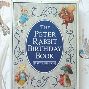 May include: A vintage "Peter Rabbit Birthday Book" featuring illustrations of Beatrix Potter characters. The cover is light grey with a navy blue oval containing the title and author's name. The book is a classic children's item.
