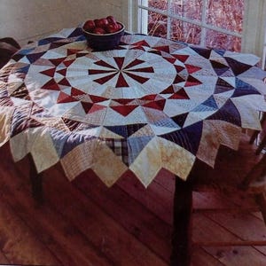 May include: A round, multi-coloured patchwork quilt tablecloth with a central starburst design. The tablecloth is laid on a wooden table with a chair beside it.