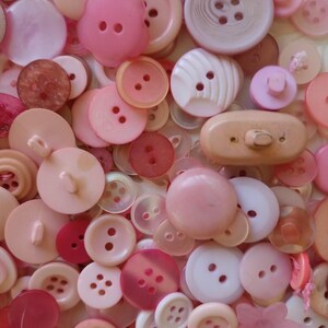 May include: A selection of pink buttons in diverse shapes and sizes. The buttons vary from pale blush to deep rose, including round, flower, and oval designs. The collection presents a range of textures and shades, with two or four-hole configurations, creating a monochromatic display.
