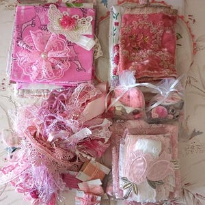 May include: A collection of craft supplies in shades of pink, including fabric squares with lace and butterfly embellishments, a piece of rose-coloured velvet, two small organza bags filled with buttons, spools of embroidery thread, and lace trim.