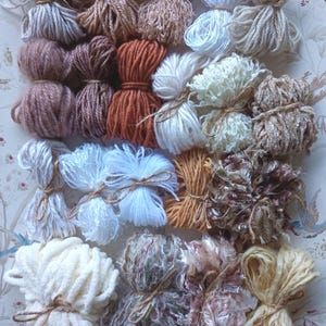 May include: A selection of yarn bundles in shades of white, beige, brown, and orange, each tied with twine. The yarn has diverse textures, from smooth to fluffy, ideal for various craft projects.