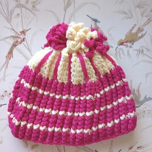 May include: A crocheted bag in fuchsia pink and cream. The bag has a ribbed texture with cream stripes and a gathered top with a large flower. The handmade bag is a unique accessory. The bag is a unique accessory.