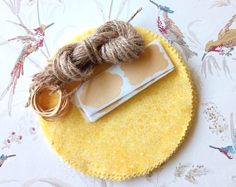 May include: A stack of circular yellow fabric pieces with pinked edges, topped with a coil of brown jute twine, a small wooden ring, and a strip of blank, scalloped-edge adhesive labels in a light tan colour.