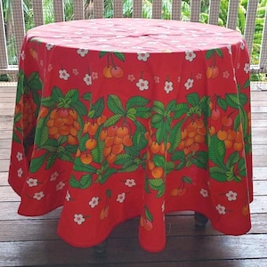May include: A round red tablecloth with a repeating pattern of cherries and white flowers. The tablecloth is draped over a small table. The cherry design features green leaves and orange cherries.