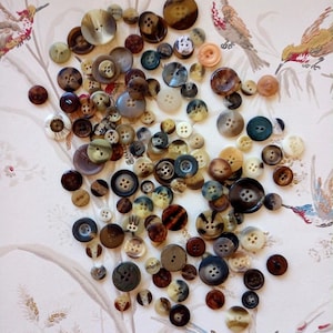 May include: A large collection of assorted round buttons in various sizes, colours, and materials, including brown, black, beige, and grey tones. The buttons feature different numbers of holes, ranging from two to four, and exhibit diverse textures and finishes.
