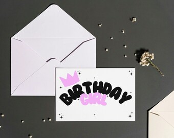 Può includere: Un biglietto bianco con una corona rosa e il testo "Birthday Girl" in lettere nere e rosa. Il biglietto è su una superficie grigia con una busta bianca e piccole perline bianche.
