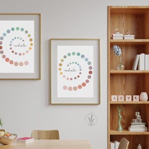 May include: Two framed prints with colorful dot spirals and the words "inhale" and "exhale" in a minimalist style. The prints are hanging on a white wall above a wooden table.