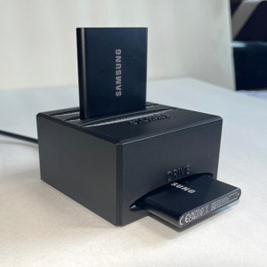May include: A black external hard drive charging station with a Samsung external hard drive and a Samsung battery inside. The station has the word "DRIVE" printed on the front.