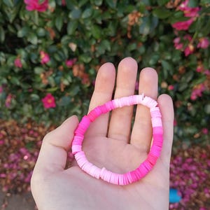 May include: A pink and white ombre bracelet made of small, round beads. The bracelet is held in a hand against a background of green leaves and pink flowers.