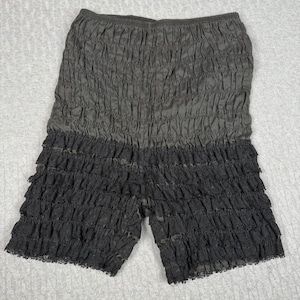 May include: Gray and black ruffled shorts. The shorts have an elastic waistband and are made of a textured fabric. The lower portion of the shorts features multiple layers of black ruffles.