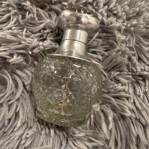 May include: A clear glass perfume bottle with a silver-colored metal top and neck. The bottle has a textured, patterned surface. The top is round with a decorative design. The bottle is sitting on a gray, fuzzy surface.