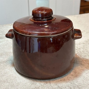 May include: A brown ceramic pot with a lid. The pot has two small handles on either side and a round knob on the lid. The pot and lid are a glossy brown color.