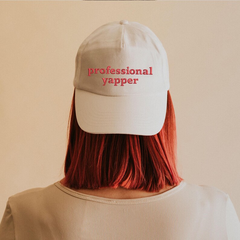 May include: White baseball cap with pink embroidered text that reads "professional yapper".