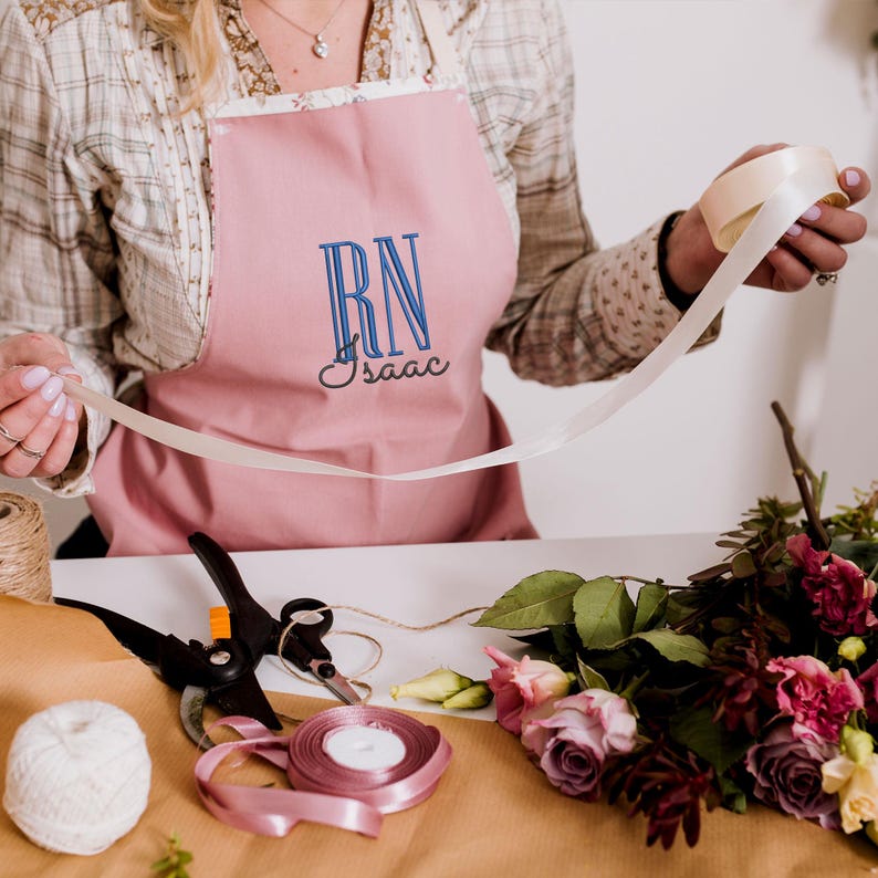 May include: A pink apron with the letters "RN" and the name "Isaac" embroidered in blue thread. The apron is being worn by a person who is holding a white ribbon.